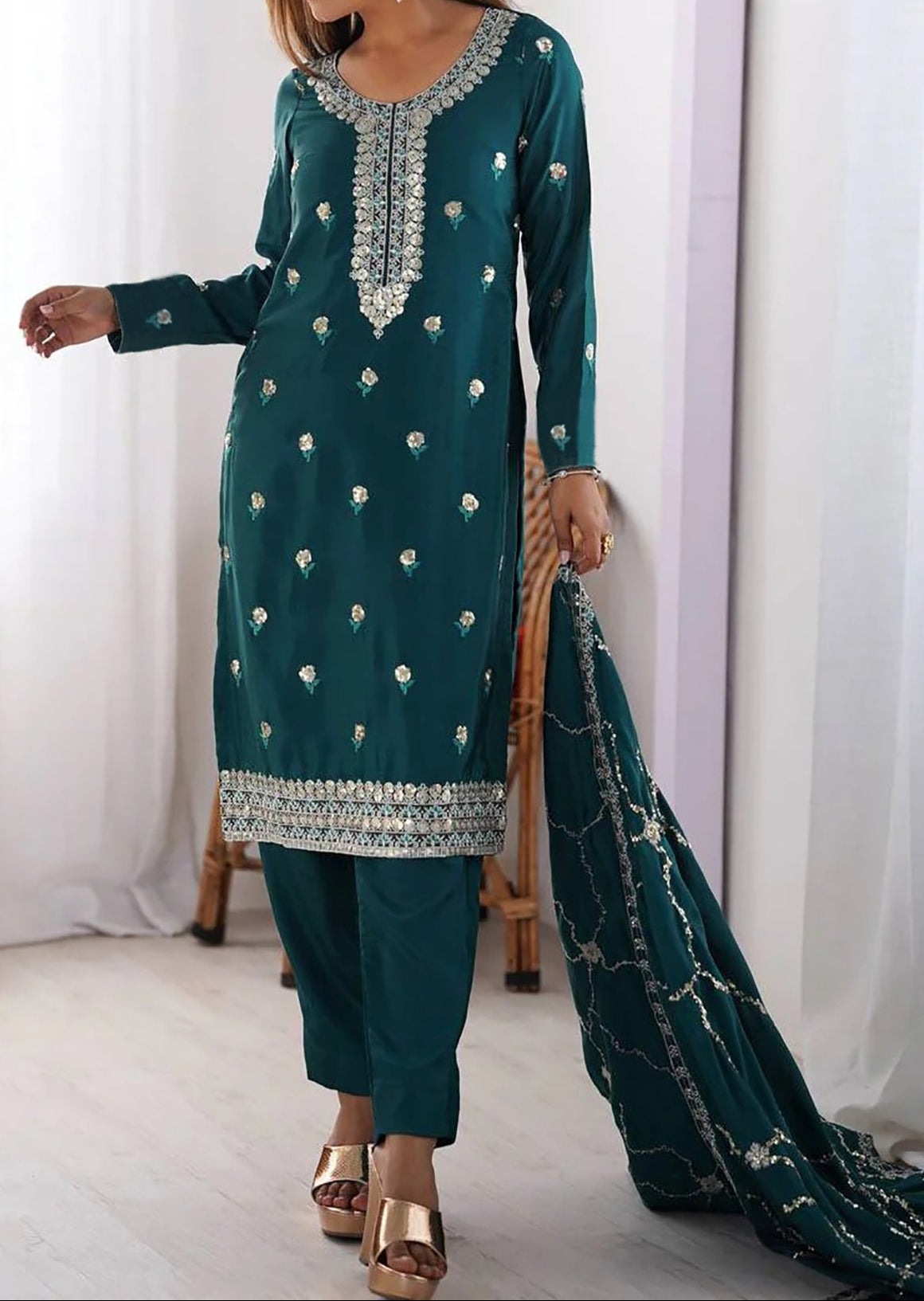 Woman wearing a teal embroidered outfit with a white curtain background